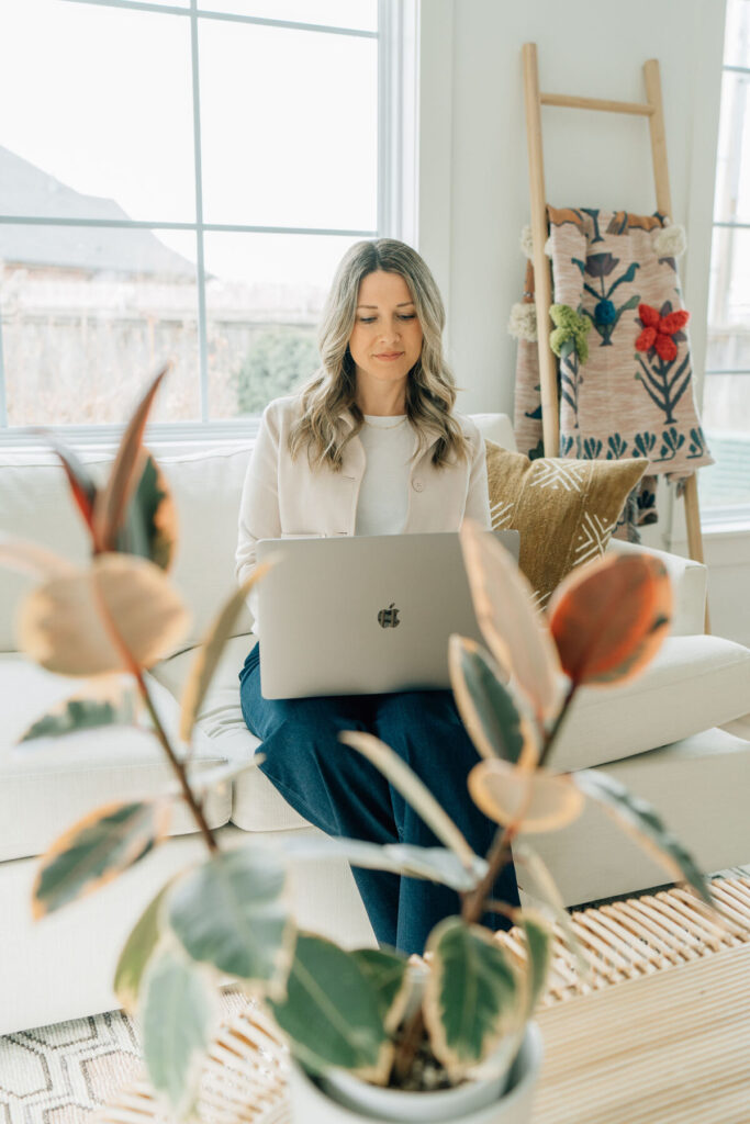 A picture of me sitting on a couch working on my laptop. There is a pretty window behind me and a plant in the foreground.